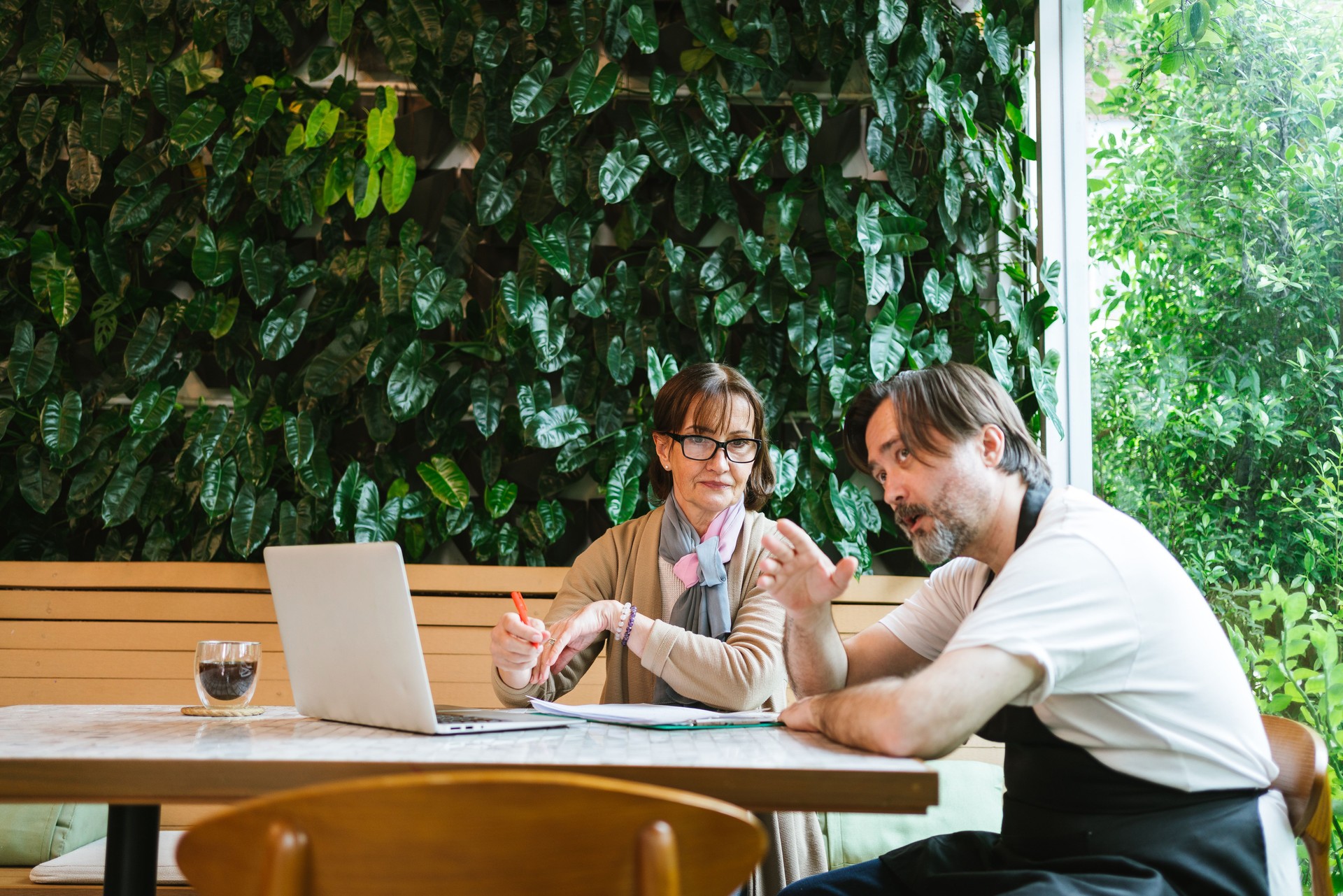 Small business cafe owner in apron meets with financial advisor in cozy green cafe, discussing strategy with laptop and notes, symbolizing diverse entrepreneurship, finance, and collaboration.